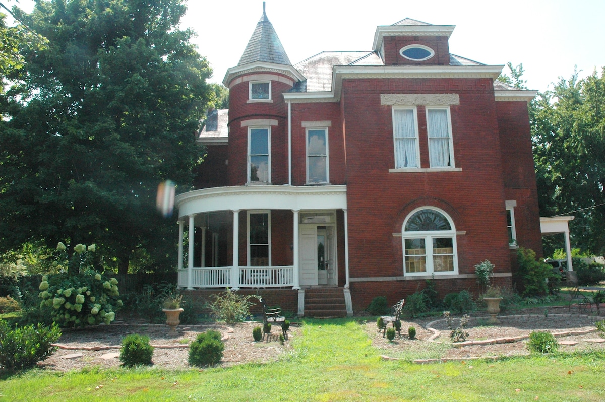 A historic red brick home is showcased, featuring a tall, pointed turret and a spacious front porch with white railings. Lush green trees and neatly trimmed garden beds surround the property, enhancing its serene outdoor setting.