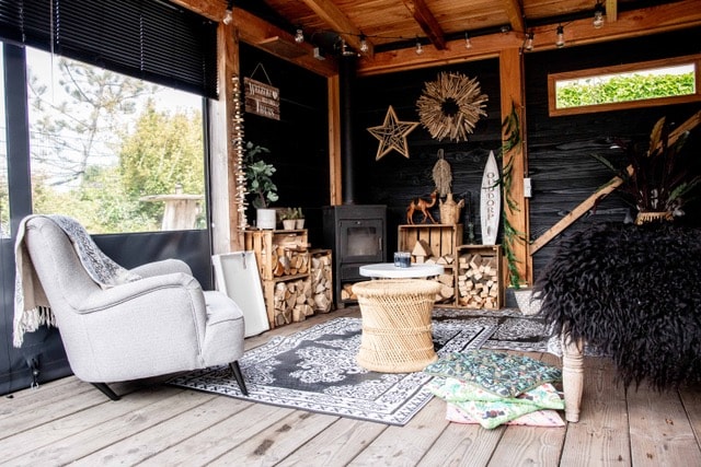 A cozy sitting area features a comfortable light armchair beside a decorative rug on wooden flooring. Shelving holds stacked firewood, and natural elements are incorporated with plants and art on the walls. A table centers the space, inviting relaxation and conversation.