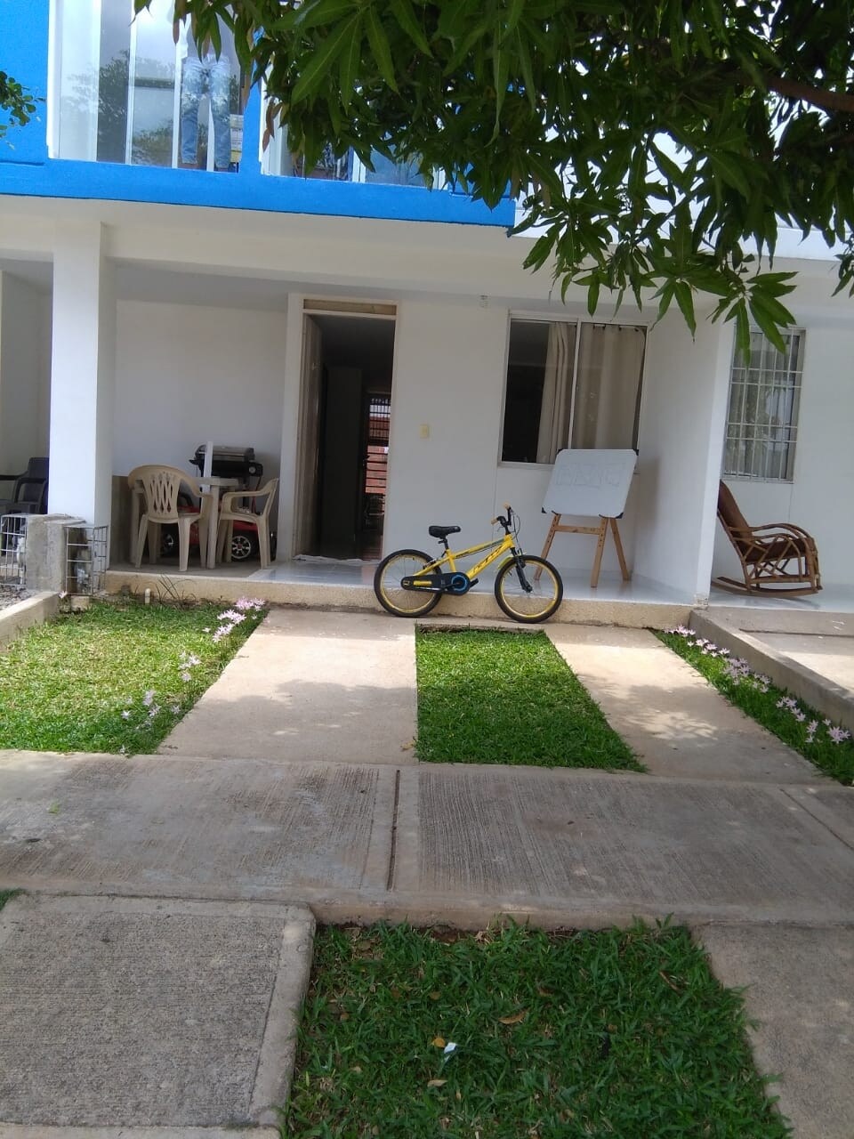 A well-maintained outdoor area features a manicured lawn with a small bicycle resting on a concrete path. The entrance to a two-story house is visible, with a set of chairs and a board nearby, framed by greenery and blue accents.