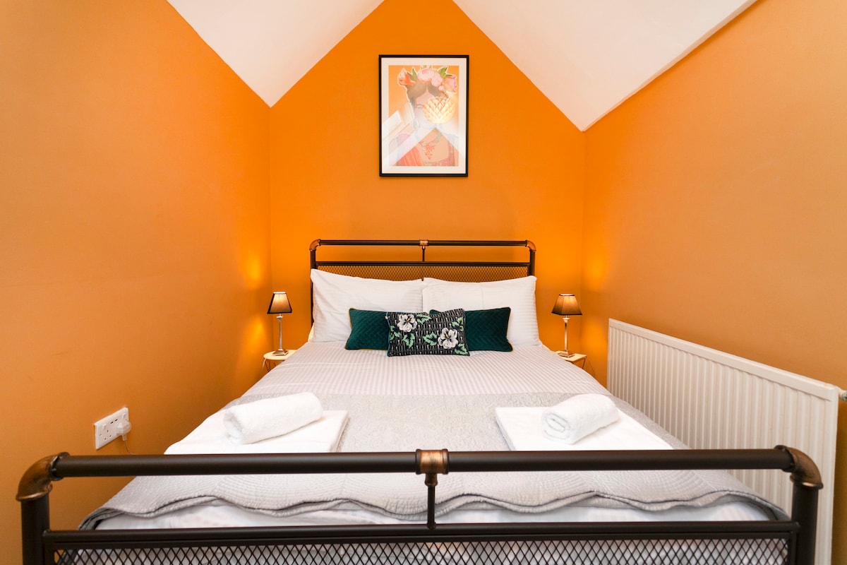 A cozy bedroom features an orange accent wall, complemented by a spacious double bed adorned with white linens. Two bedside lamps provide soft lighting, and a decorative piece hangs above the bed, enhancing the room's character. A radiator is positioned nearby.