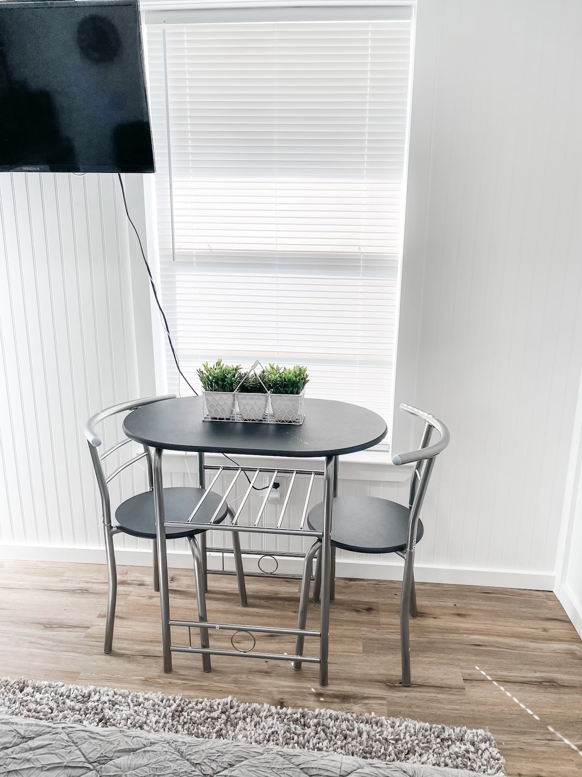 A small dining area is visible, featuring a round table with two chairs. A minimalistic centerpiece of potted plants is placed on the table. Natural light enters through a nearby window adorned with blinds.