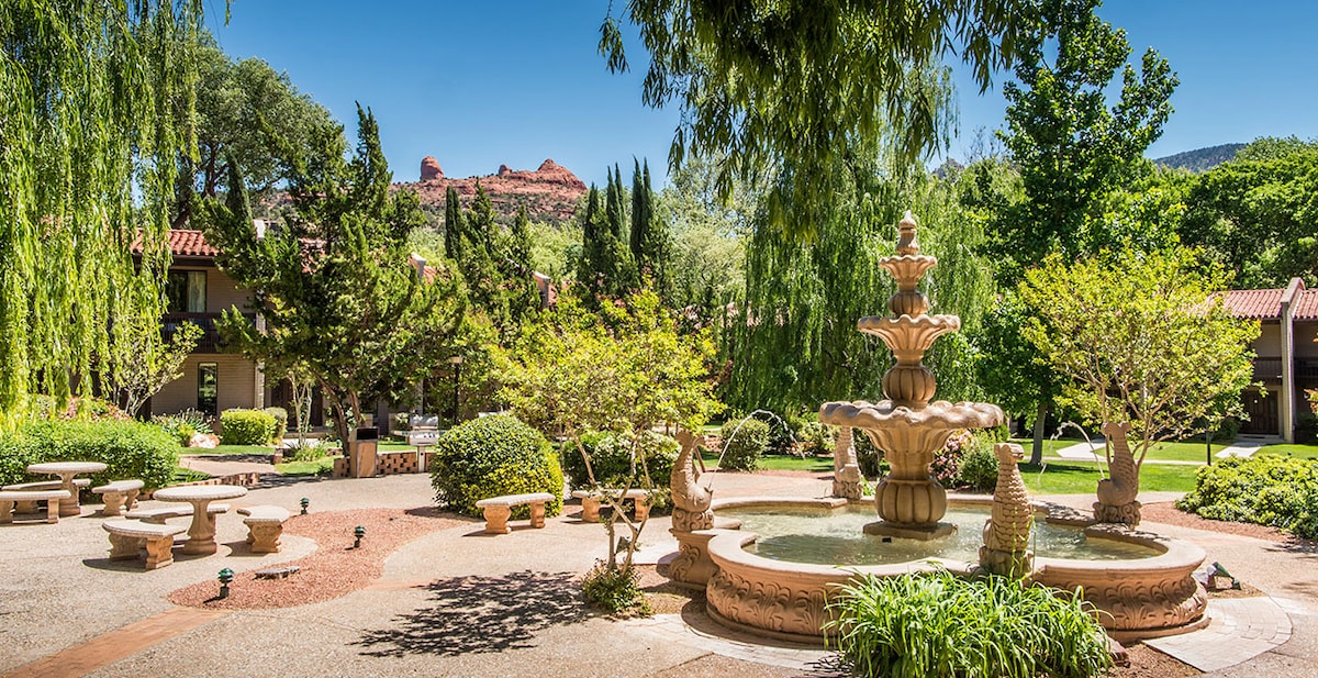 A landscaped courtyard features a central fountain surrounded by decorative stone benches and lush greenery. The backdrop includes trees and vibrant foliage, with red rock formations visible in the distance under a clear blue sky.