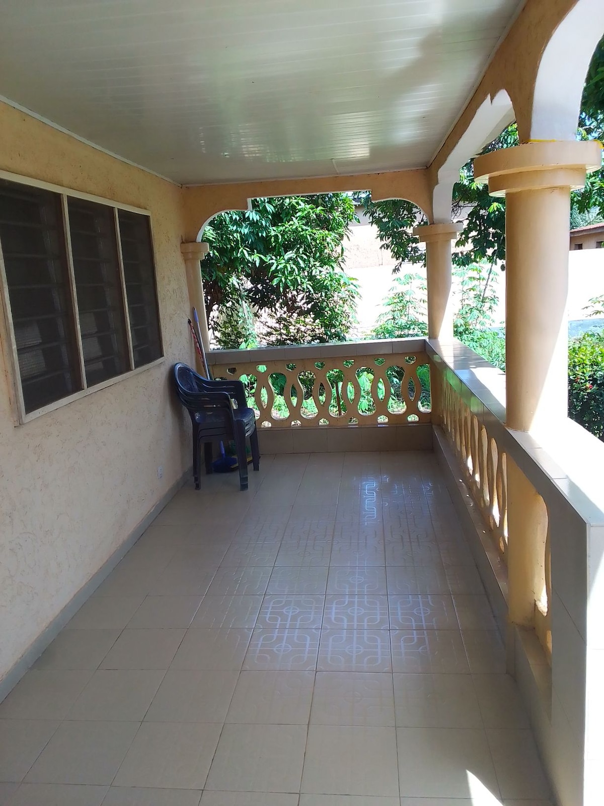 A covered terrace is visible, featuring a tiled floor and an open railing design. Two windows allow natural light to enter. A single plastic chair is placed against the wall, overlooking lush greenery, creating a serene outdoor space.