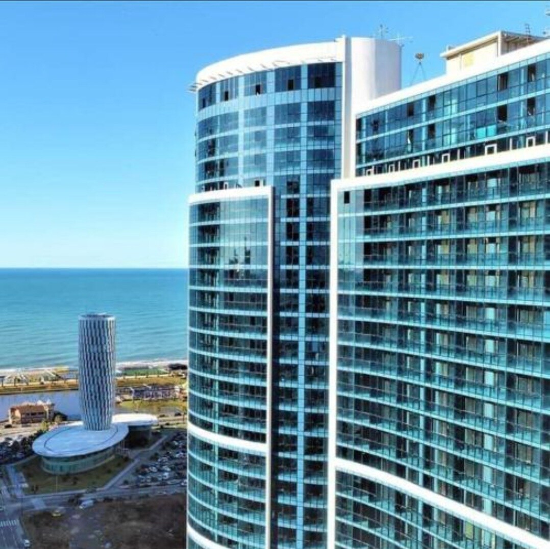 A contemporary high-rise building showcases sleek glass windows reflecting the blue sky and the sea. The structure stands prominently along the coastline, with the ocean visible in the background. The vibrant shoreline can be seen in the distance, highlighting the building's prime location.