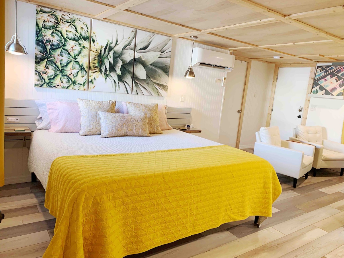 A spacious cottage interior features a large white bed covered by a bright yellow blanket. Pastel-colored pillows add subtle contrast. The wall above the bed showcases vibrant pineapple artwork, while the room is illuminated by modern light fixtures. A cozy seating area is visible to the right.