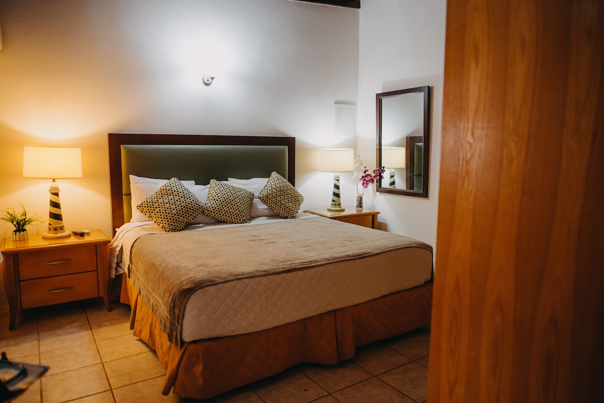 A comfortable sleeping area features a spacious bed with neutral bedding and decorative throw pillows. Two nightstands with lamps are positioned on either side. A mirror reflects the room's soft lighting, while a door leads to the adjacent space.