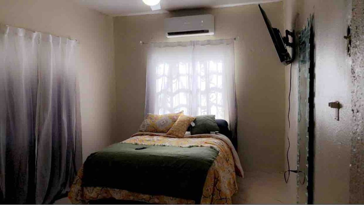 A cozy bedroom is presented with a spacious bed dressed in soft bedding. A wall-mounted television is positioned across from the bed. Natural light filters through sheer curtains, while an air conditioning unit provides climate control. Light-colored walls create an open and inviting feel.