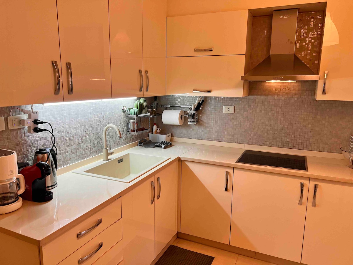 A modern kitchen features sleek white cabinetry and a neutral backsplash. A stainless steel range hood is mounted above the cooktop, while a spacious sink is positioned near counter appliances. Soft under-cabinet lighting enhances the workspace.