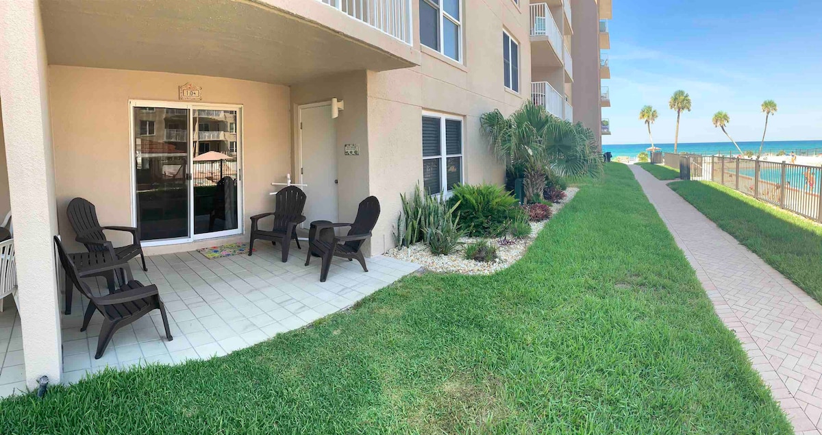An inviting patio space features several dark wood chairs arranged on tile flooring, surrounded by lush greenery and a well-maintained lawn. The patio offers views of the ocean in the distance, with a pathway leading towards the beach in the background.