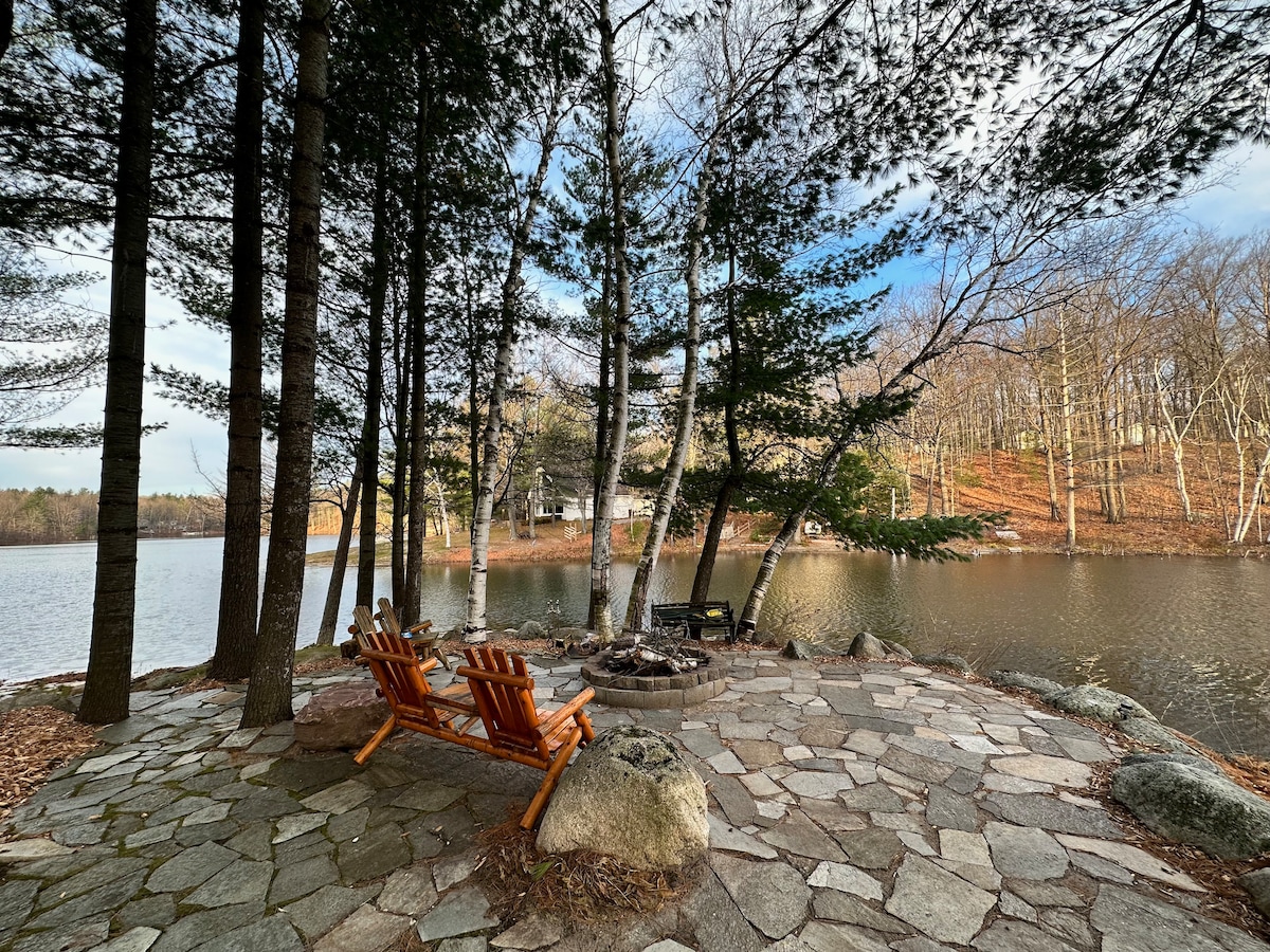A serene lakeside area features two wooden adirondack chairs positioned on a stone patio. Surrounding trees provide a natural backdrop, and a fire pit is located nearby. The calm water reflects the landscape, offering a tranquil setting for relaxation.