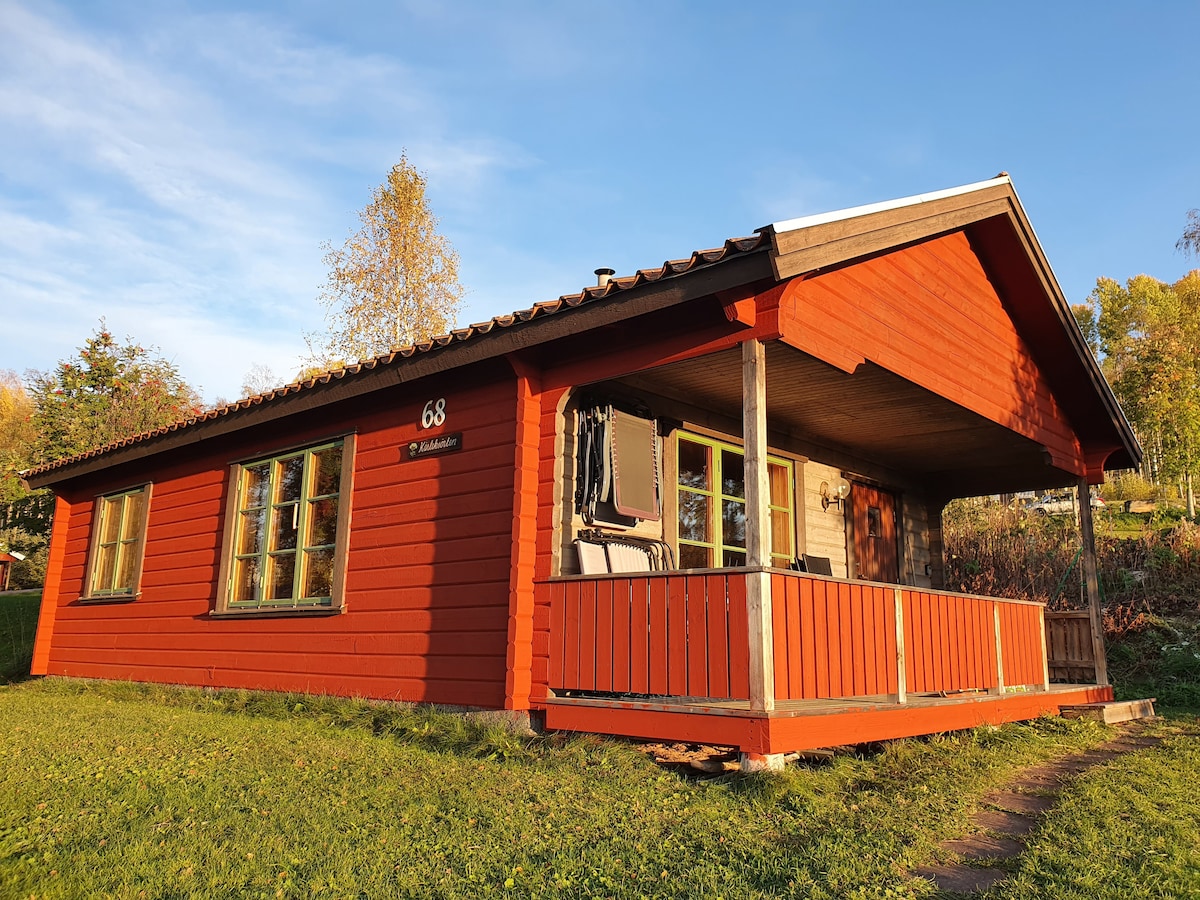 A charming red cabin is featured with a covered porch, offering multiple windows that allow natural light to enter. The surrounding greenery complements the setting, while steps lead up to the porch, enhancing accessibility to this inviting space in a tranquil area.