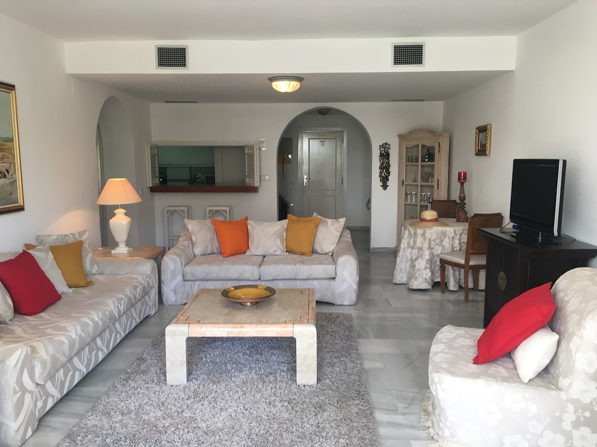 A spacious living area features two large sofas with colorful cushions and a decorative rug. A small table is set with a bowl, while a television stands on a cabinet. The room is enhanced by soft lighting from an elegant lamp and displays a bright and airy ambiance.