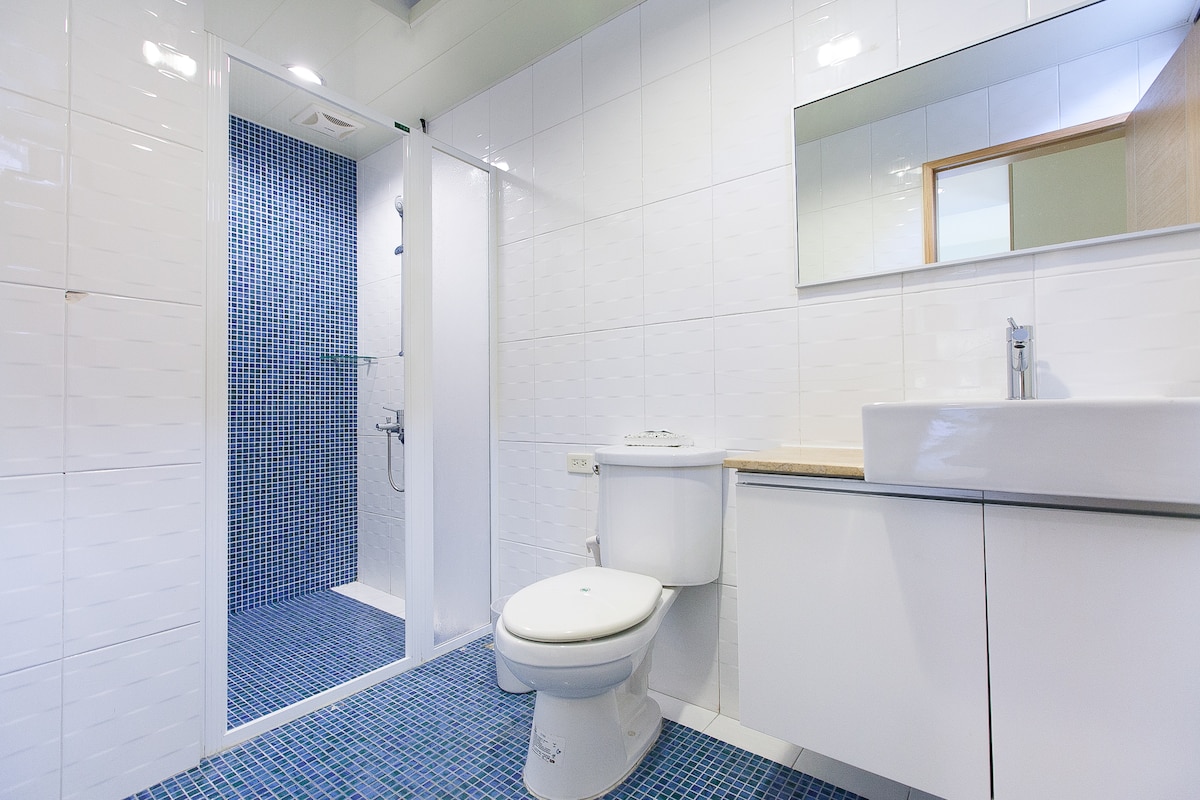 A modern bathroom features white tiled walls and a striking blue mosaic floor. A glass shower enclosure is visible, along with a sleek white sink and cabinet. A large mirror reflects the spacious layout, contributing to the room's clean and bright aesthetic.