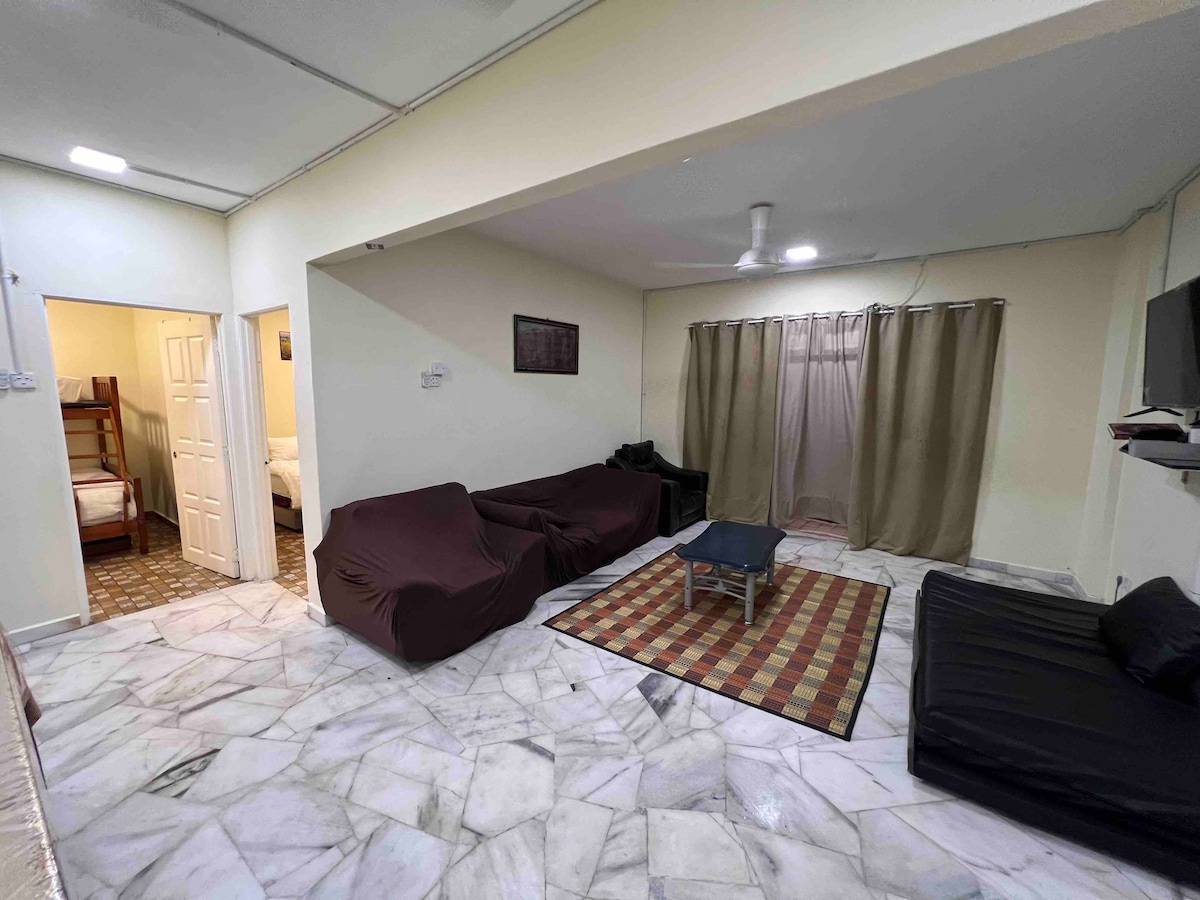 A spacious living area features a patterned area rug positioned under a low coffee table. Two covered sofas are arranged for comfort, while a ceiling fan provides ventilation. Light enters through curtained windows, with an opening leading to additional rooms visible in the background.