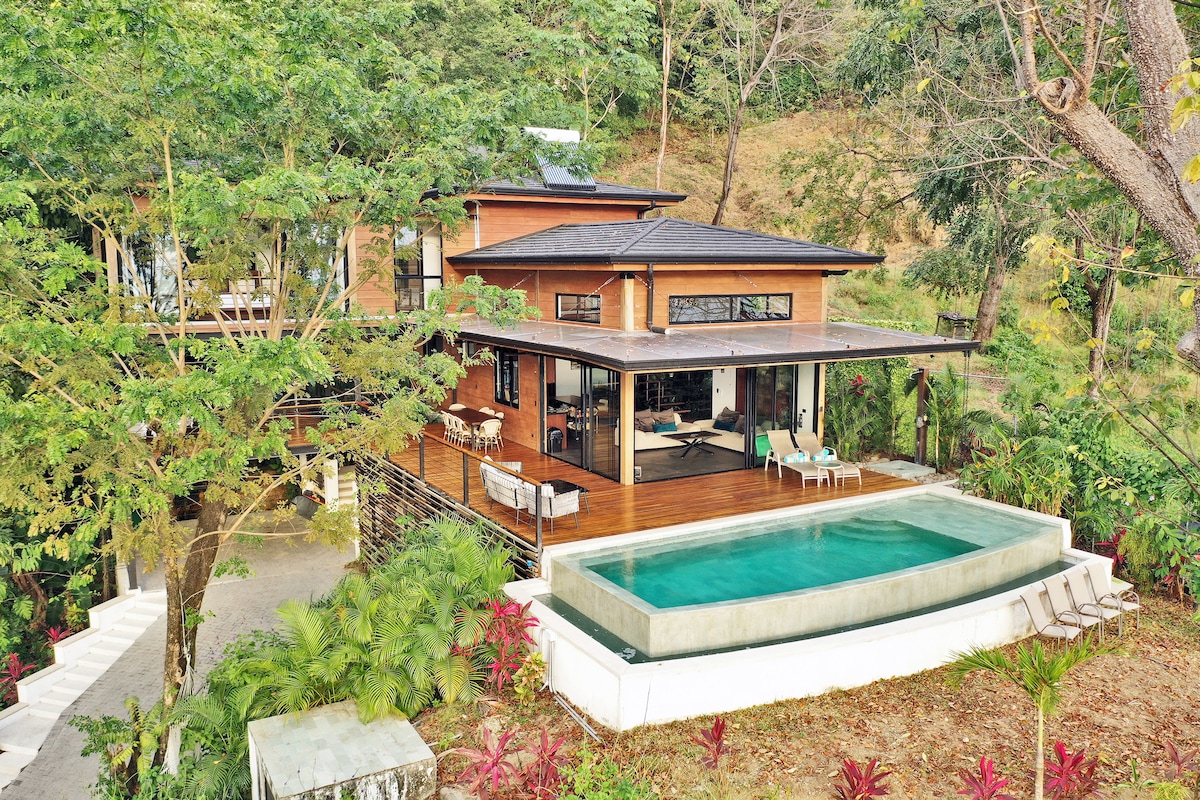The villa, set among lush greenery, features a wooden deck with outdoor seating and a private pool. Large glass windows provide views of the indoor living area, seamlessly blending with the surrounding natural landscape. Tropical plants frame the space, enhancing its tranquil ambiance.