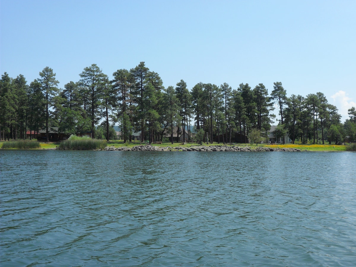 A serene lakefront view features calm waters reflecting a clear blue sky. Pine trees line the shore, with a small rocky beachfront visible. A vintage log cabin can be seen in the background, surrounded by greenery, contributing to a tranquil natural environment.