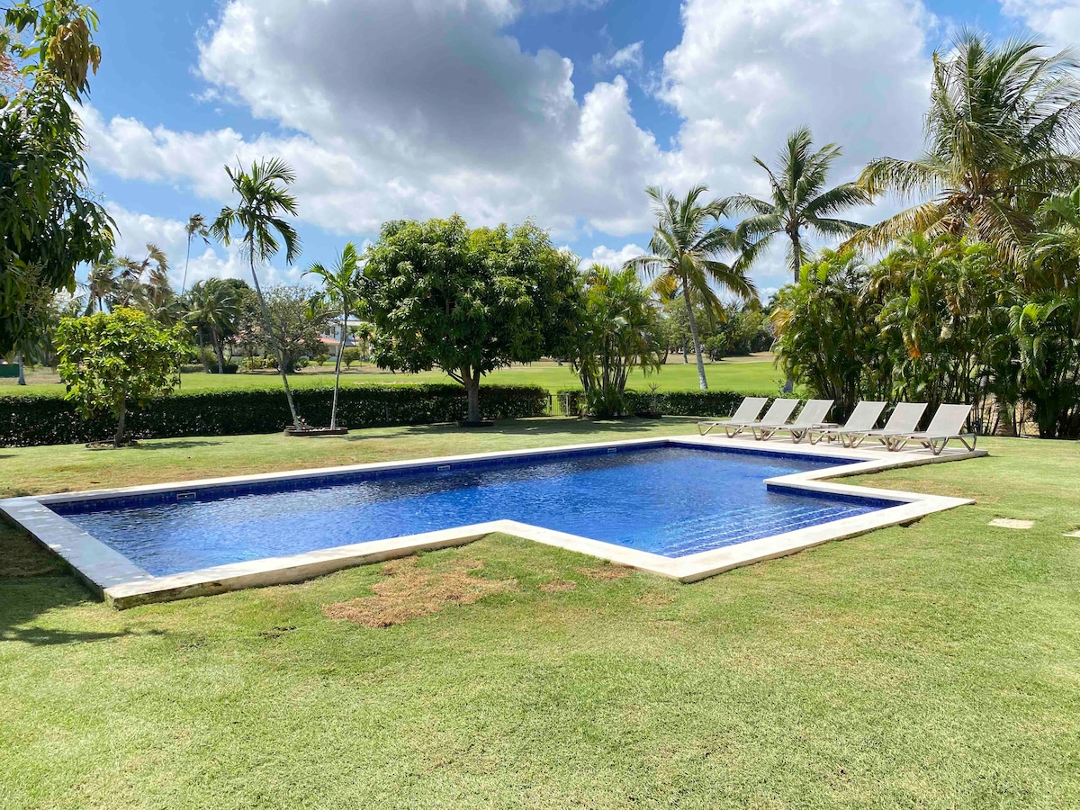 A private pool is situated within a lush tropical garden, surrounded by green grass and palm trees. Several lounge chairs are arranged neatly along one side of the pool, offering a comfortable spot to relax. The clear blue water reflects the sky and nearby trees.