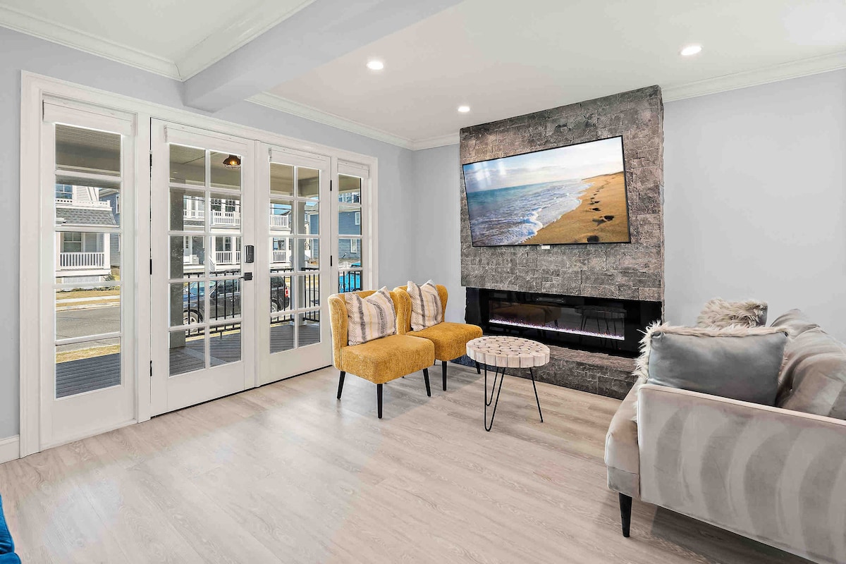 The living area showcases a cozy seating arrangement with two yellow chairs and a gray sofa. A modern coffee table is positioned in the center. A sleek fireplace and large wall-mounted TV are featured, with natural light entering through glass doors leading to the porch.