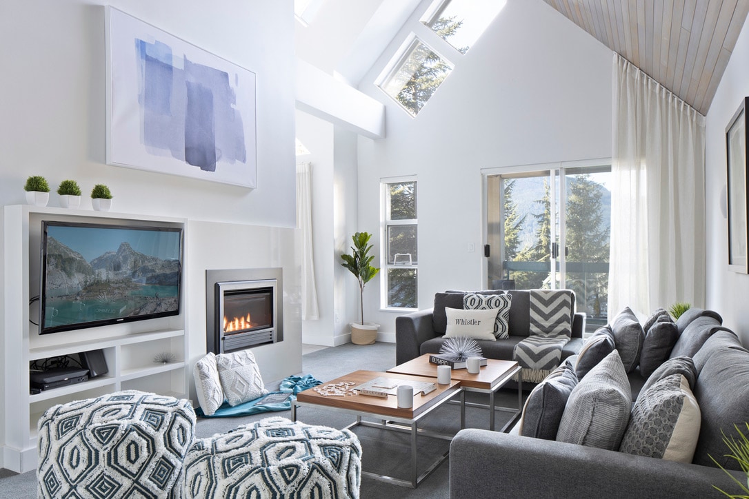 A bright living room is illuminated by natural light from large windows, highlighting a cozy gas fireplace. The space features a large sectional sofa and multiple seating options, complemented by a smart TV and decorative accents. Mountain views are visible through the balcony doors.
