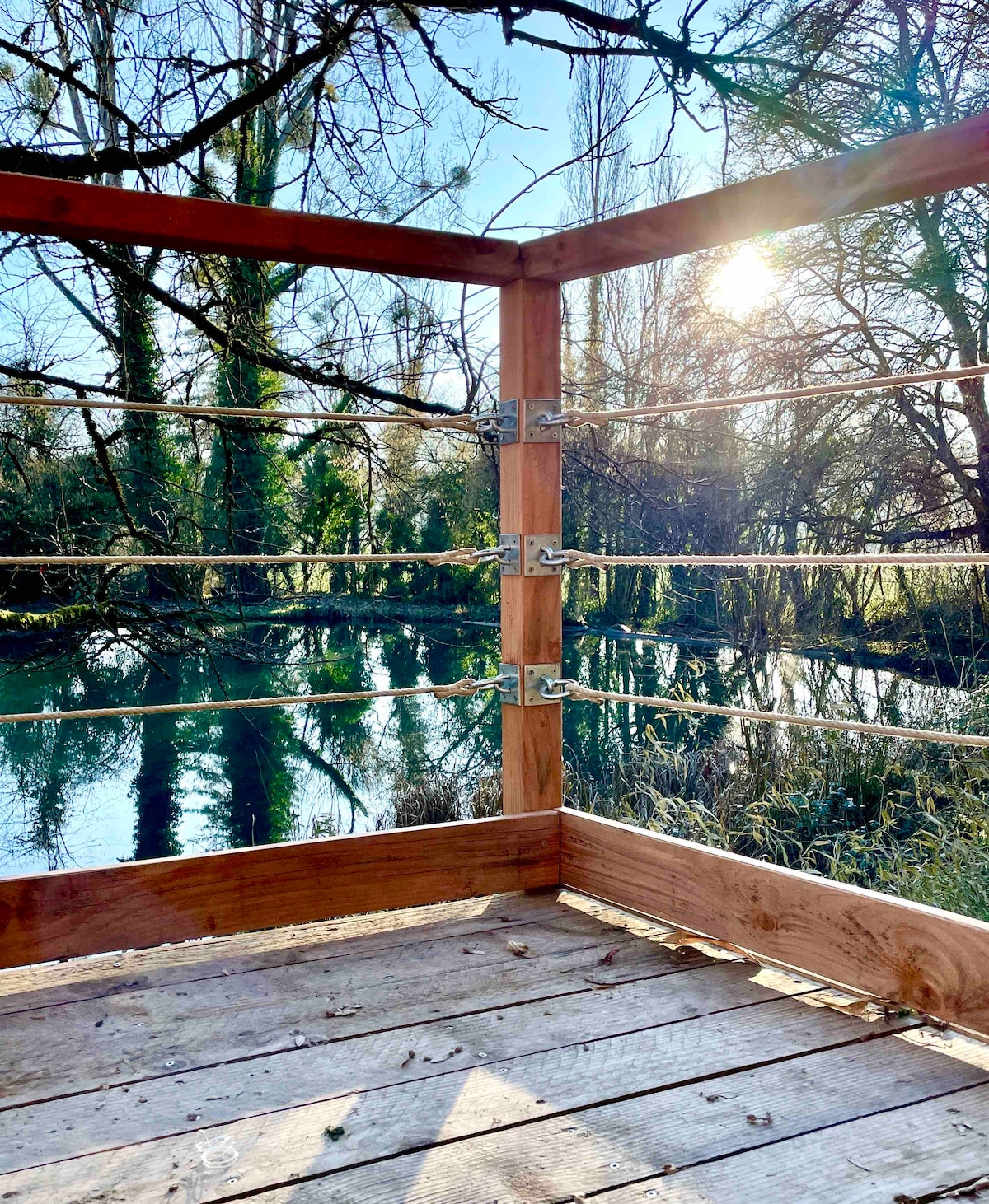 A wooden deck features a railing made of metal cables, providing a clear view of the serene water and surrounding trees. Sunlight filters through the branches, casting gentle reflections on the water's surface.