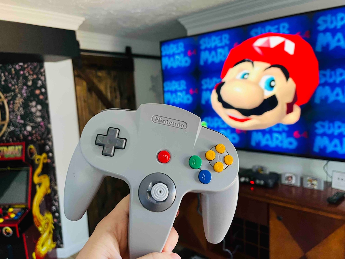 A classic gaming controller is held in front of a large TV displaying a colorful image of a character from a vintage video game. The background features a dark, patterned wall that adds a playful touch to the gaming area.