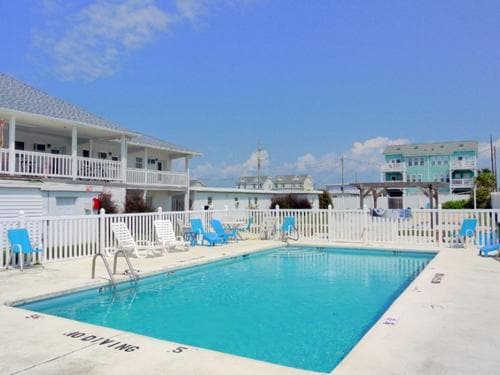A clear blue sky reflects over a swimming pool surrounded by a white fence. Lounge chairs are placed around the pool area, and a second building is visible in the background, adding to the inviting atmosphere.