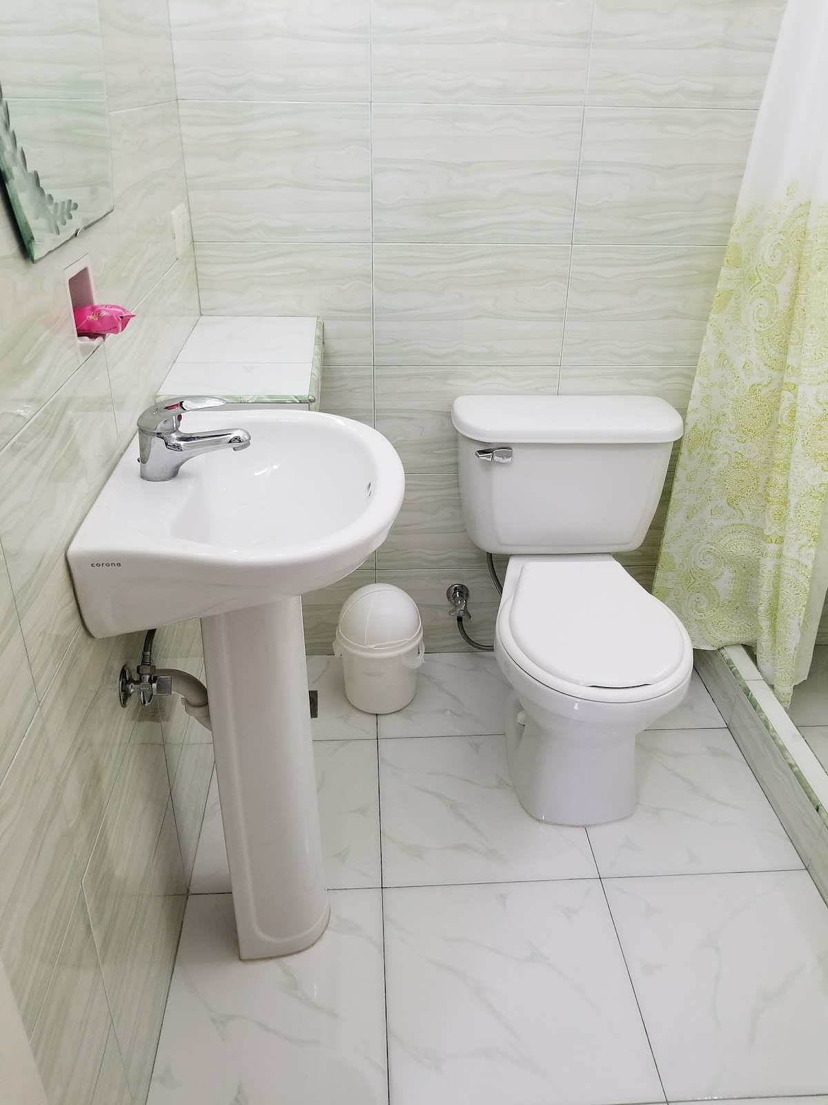 The bathroom is equipped with a pedestal sink and a modern toilet. A white shower curtain with a subtle green pattern adds a touch of color. White tiles line the floor and walls, contributing to a bright and clean atmosphere.