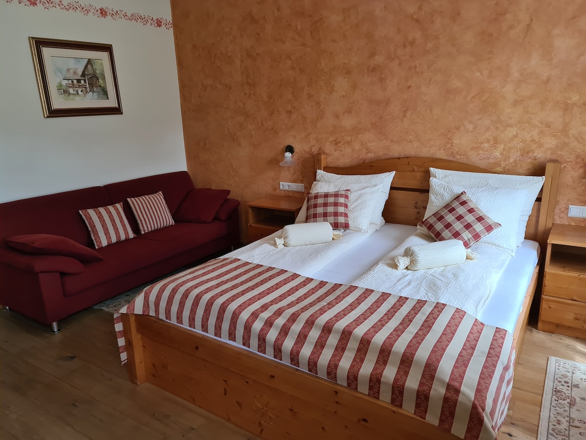 A cozy sleeping area features a spacious bed with striped bedding and plaid accent pillows. A soft red couch complements the room's warm tones, while a wooden nightstand and decorative wall art add subtle charm.