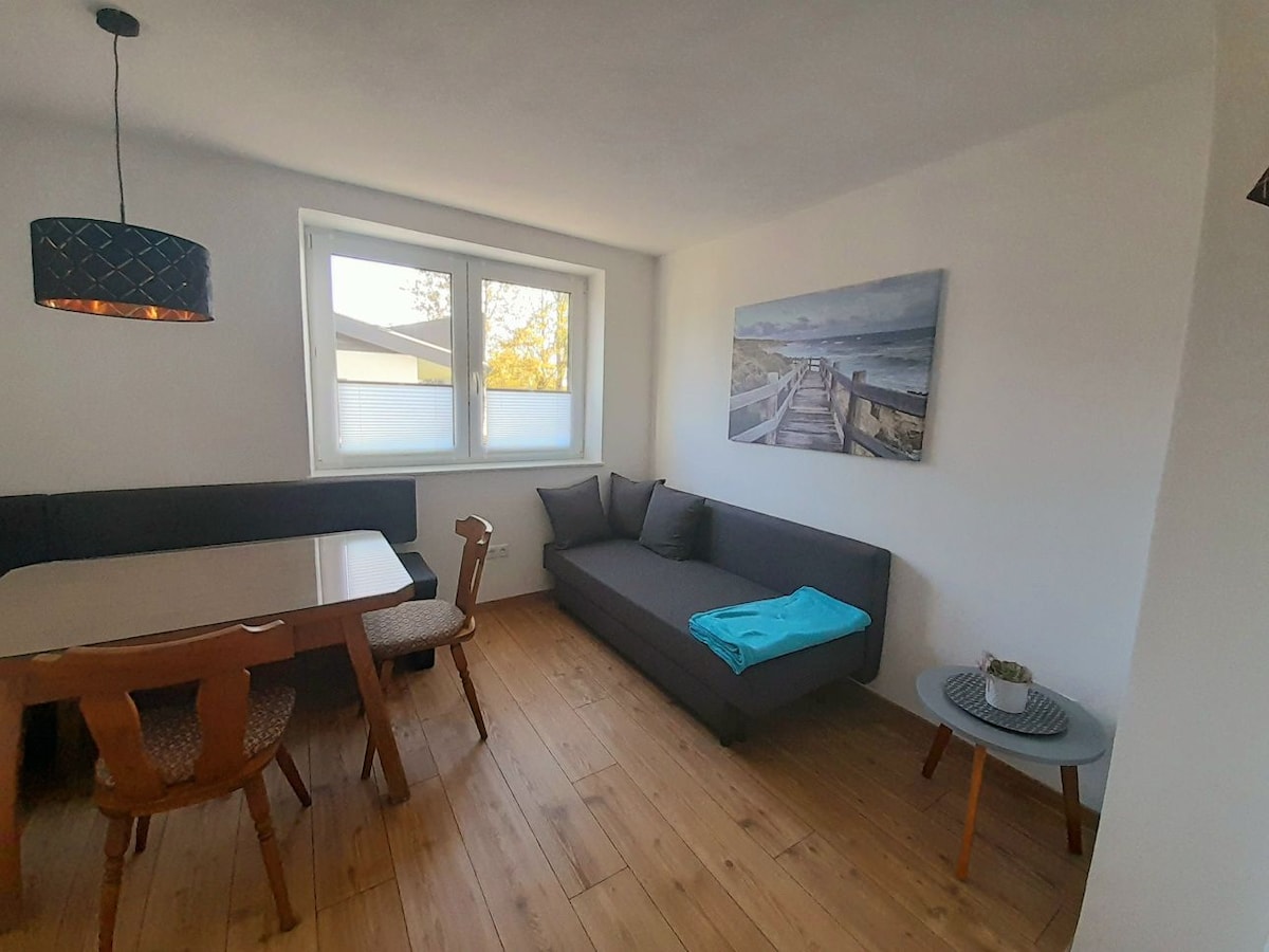 A well-lit living and dining area features a dining table with four wooden chairs and a comfortable gray couch, complemented by a small side table. Large windows allow natural light to illuminate the space, while a serene beach-themed painting adds visual interest.