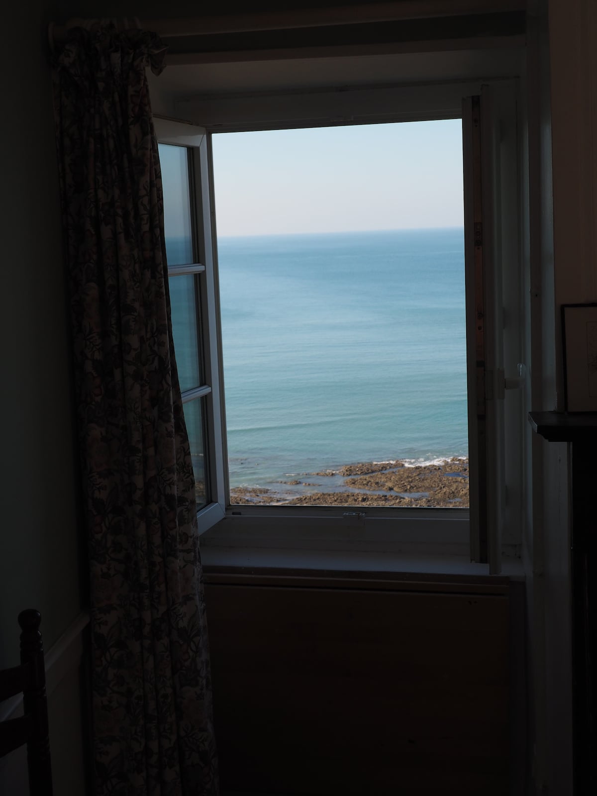 A window is framed by patterned curtains, revealing a serene view of the sea and rocky shoreline. The water's surface reflects soft hues of blue, while the distant horizon meets the sky, creating a tranquil coastal scene.