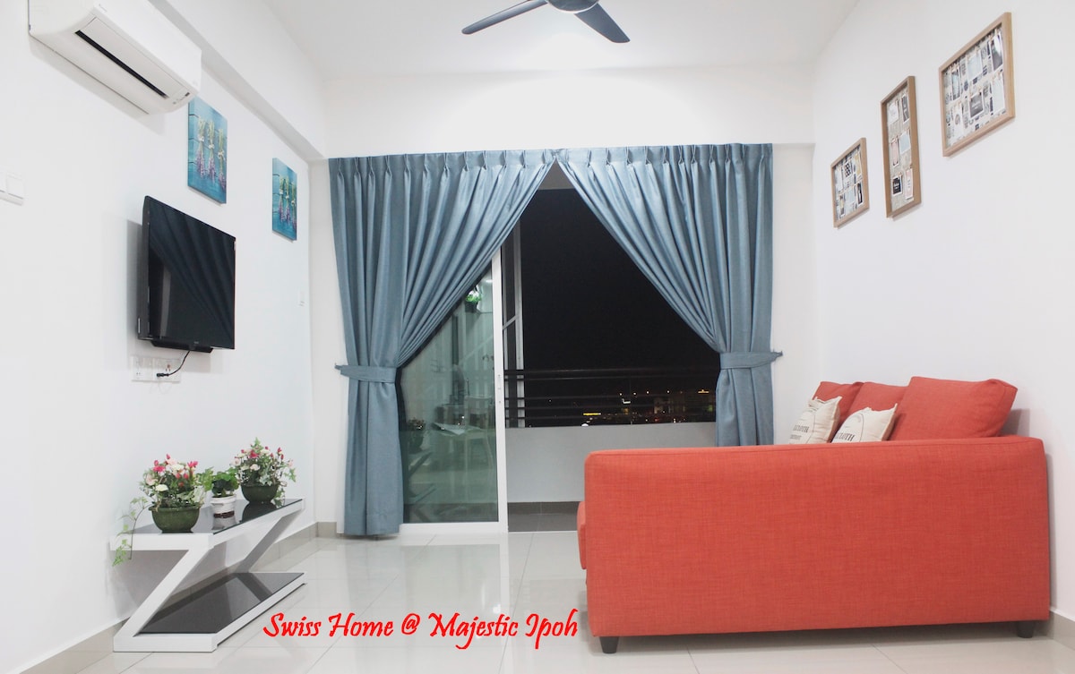 A modern living area features a bright orange sofa positioned against a white wall. Floor-to-ceiling curtains frame a sliding glass door that opens to a balcony. A television is mounted on the wall, and small potted plants add a touch of greenery to the space.
