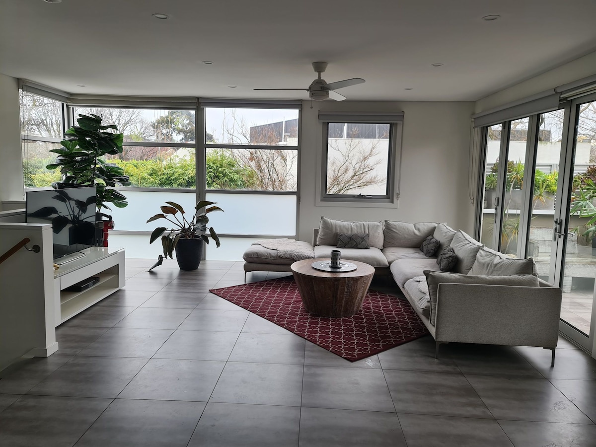 A bright living area features large windows allowing natural light. A spacious sectional sofa is centered around a wooden coffee table, with a decorative plant positioned in the corner. A TV is mounted on the wall, creating a comfortable space for relaxation.