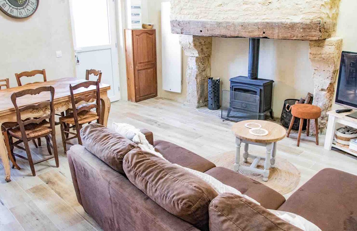 A cozy living area features a comfortable sofa positioned near a wood-burning stove set within a rustic stone fireplace. A round coffee table rests on a circular rug, while a dining table with wooden chairs is placed nearby. Natural light flows through a window, enhancing the inviting space.