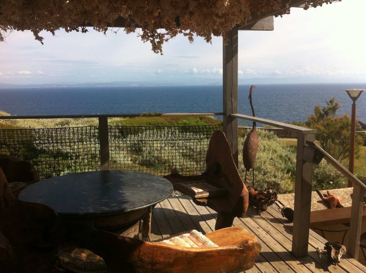 An outdoor terrace is shown, featuring unique wooden seating and a round table. The space is shaded by decorative overhead elements, offering a panoramic view of the sea and the distant shoreline. Lush greenery is visible in the foreground, enhancing the natural setting.