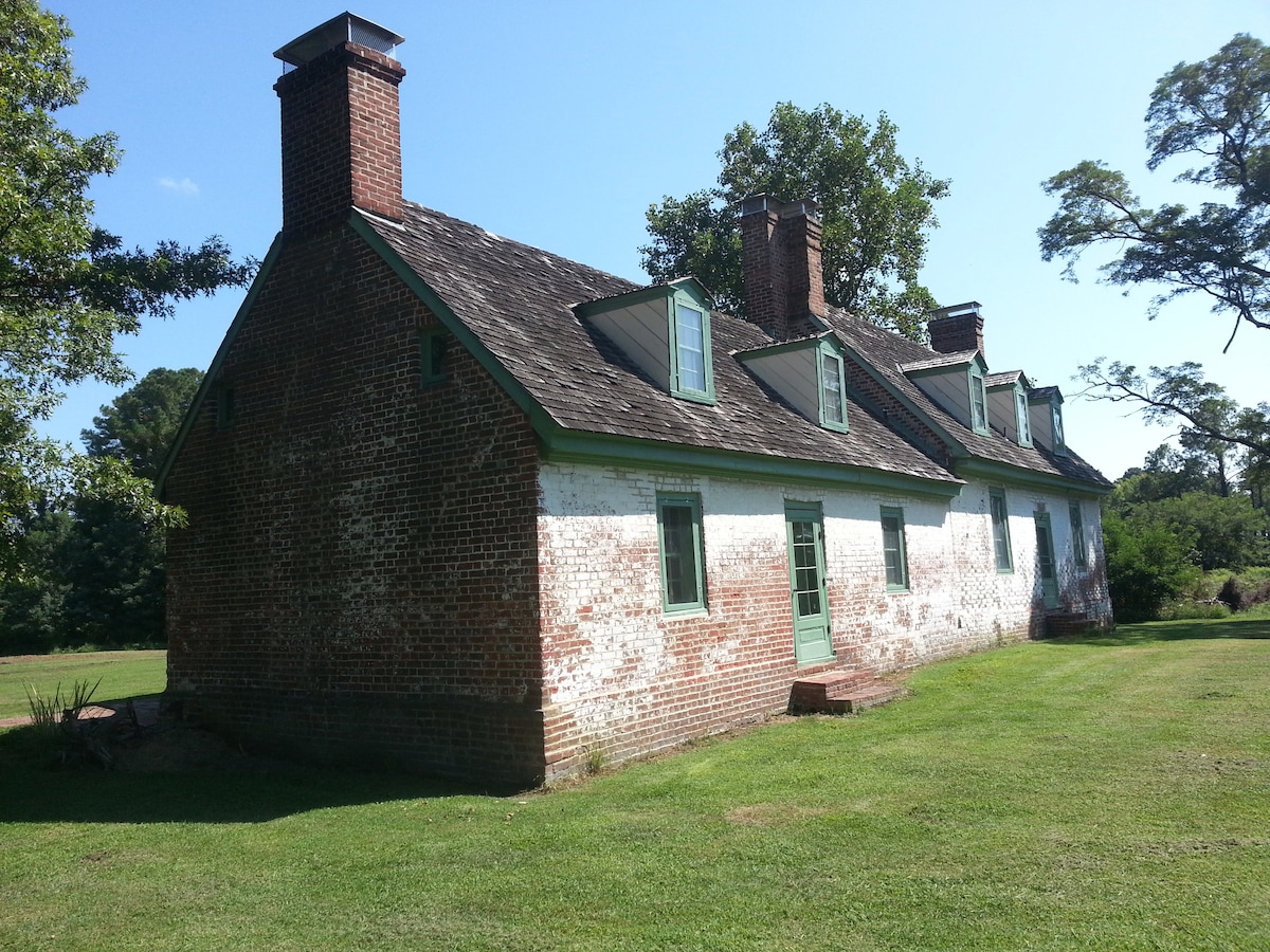 The side view of a historic brick house is shown, featuring a gabled roof with multiple chimneys. The whitewashed brick exterior contrasts with the green trim around the windows and door. The well-maintained lawn surrounds the structure, complemented by mature trees in the background.