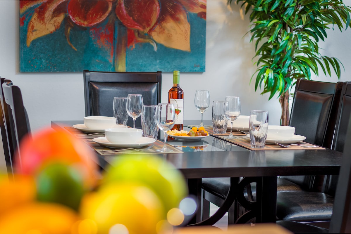 A formal dining table is set with dinnerware and glassware, surrounded by black chairs. A centerpiece of fruits is displayed, complementing the vibrant floral artwork hung on the wall. Soft green foliage adds a fresh element to the setting.
