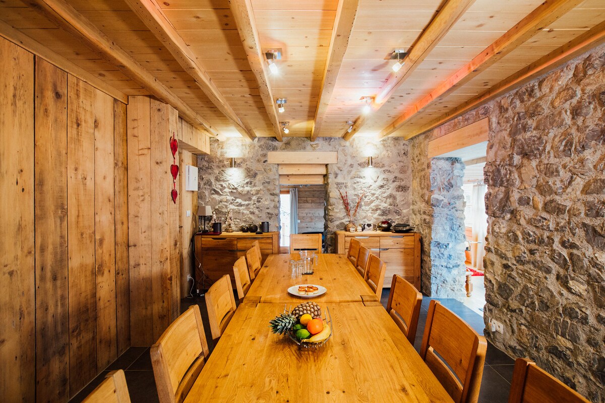 The dining area features a long wooden table surrounded by multiple wooden chairs, accommodating up to 15 guests. Exposed wooden beams and stone walls create a rustic ambiance, while soft lighting highlights the natural materials.
