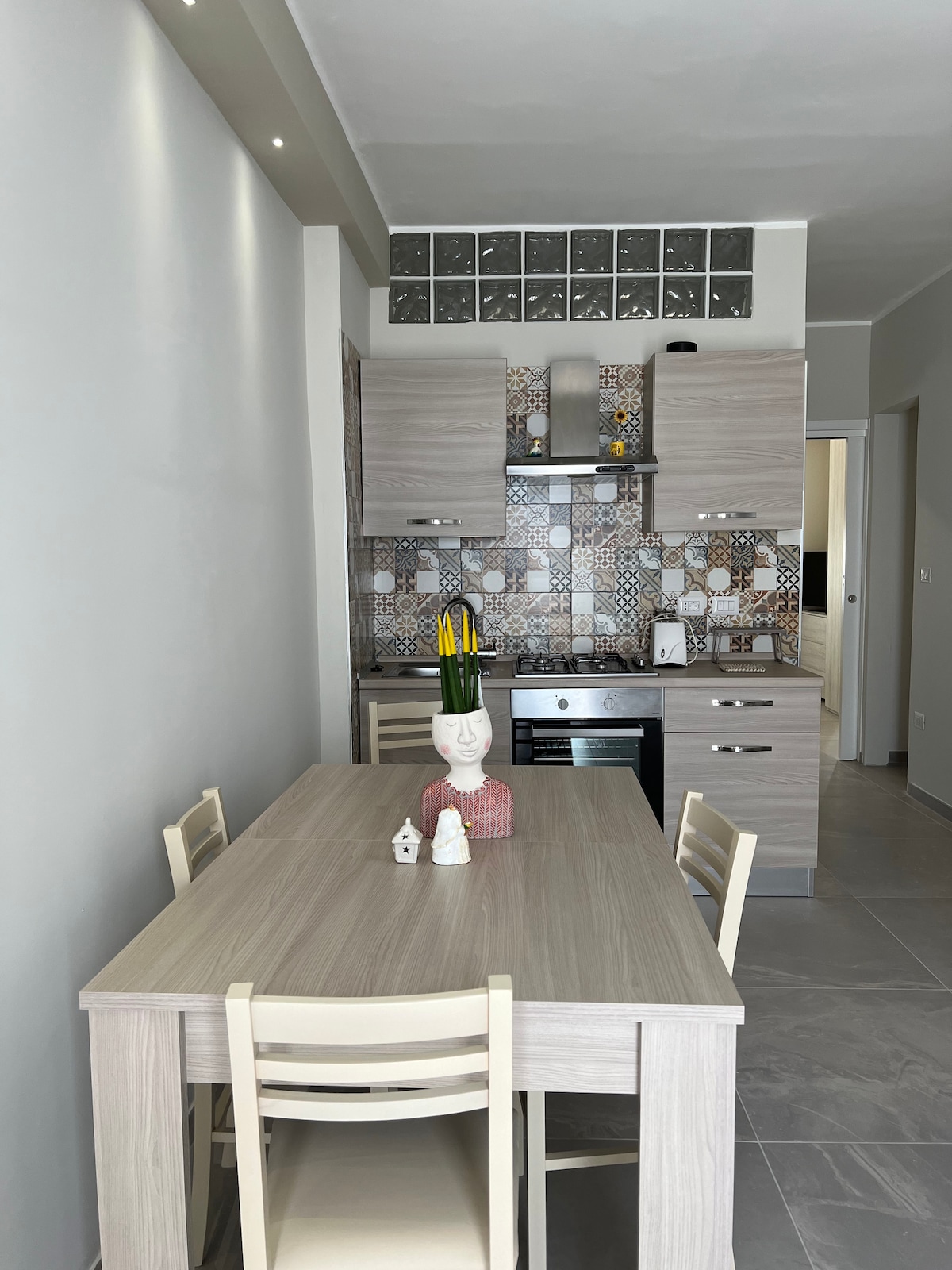 A modern kitchen area features light wooden cabinetry and a stylish backsplash with geometric patterns. A simple dining table with four chairs occupies the space, complemented by a decorative figure as a centerpiece. Natural light brightens the room, highlighting its clean lines and contemporary design.