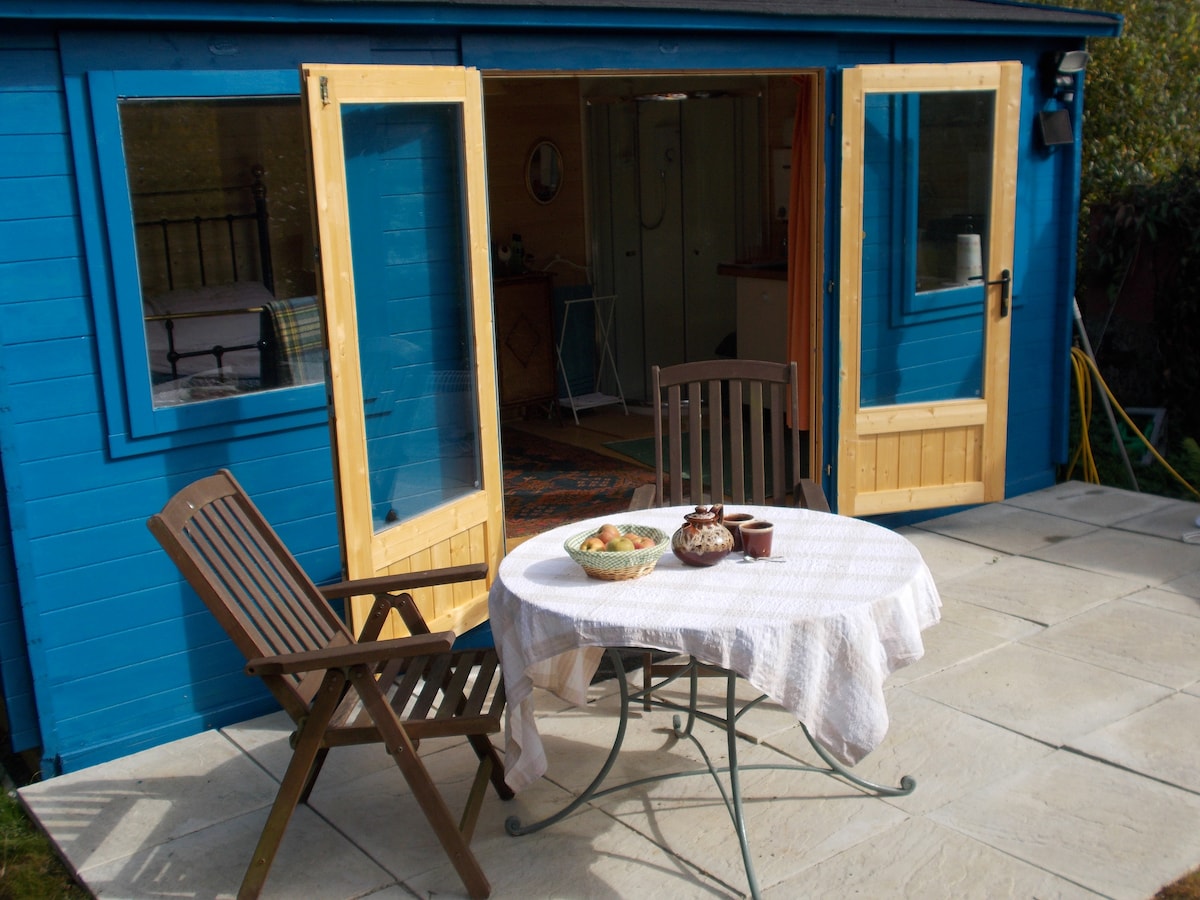 An outdoor space features a round table with a white tablecloth, accompanied by a bowl of fruit and a traditional teapot. Two wooden chairs are positioned beside the table. Large double doors open into a compact cabin with pastel blue walls, providing an inviting entryway.