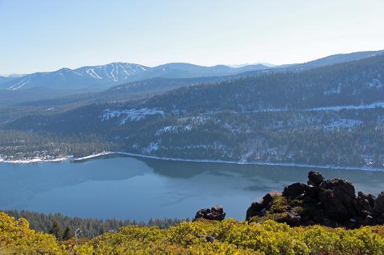 A panoramic view of a serene lake is framed by rolling mountains under a clear sky. The tranquil water reflects the surrounding greenery and rocky terrain, while patches of snow hint at a cooler climate. The scene conveys a sense of natural beauty and calm.