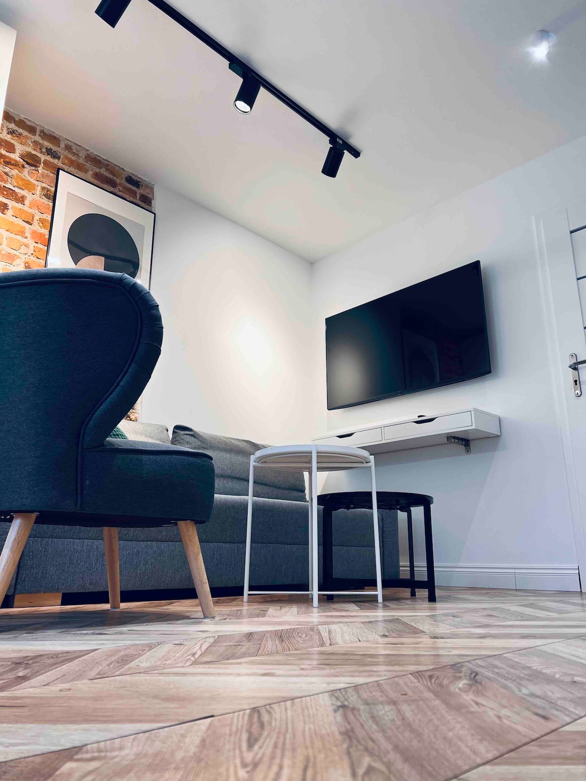 A cozy living area features a combination of seating options, including a plush armchair and a gray sofa. A flat-screen television is mounted on the wall, with a small table positioned nearby. Light from recessed ceiling fixtures highlights the warm wood-patterned flooring.