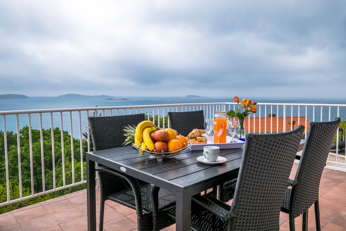 A dining table set for a meal is shown on a terrace, overlooking the sea. A fruit bowl filled with bananas, oranges, and pineapples is centrally placed, accompanied by glasses and a cup. The distant coastline can be seen under a cloudy sky.