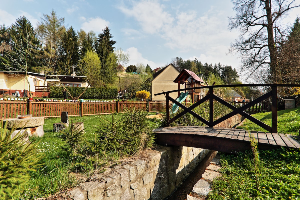 A wooden footbridge crosses a small creek, leading into a green yard featuring a play structure. The area is surrounded by gentle hills and trees, with well-maintained grass and shrubbery. In the background, residential buildings are visible, blending with the natural landscape.
