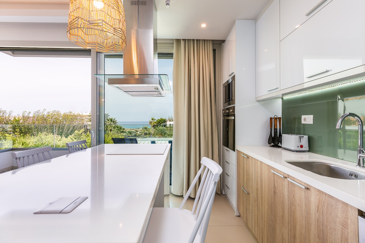 A modern kitchen space features sleek white cabinetry and wooden accents. A long kitchen island with minimalist white chairs provides seating. Natural light flows in through large windows, revealing a glimpse of the sea view beyond. The space is equipped with contemporary appliances.