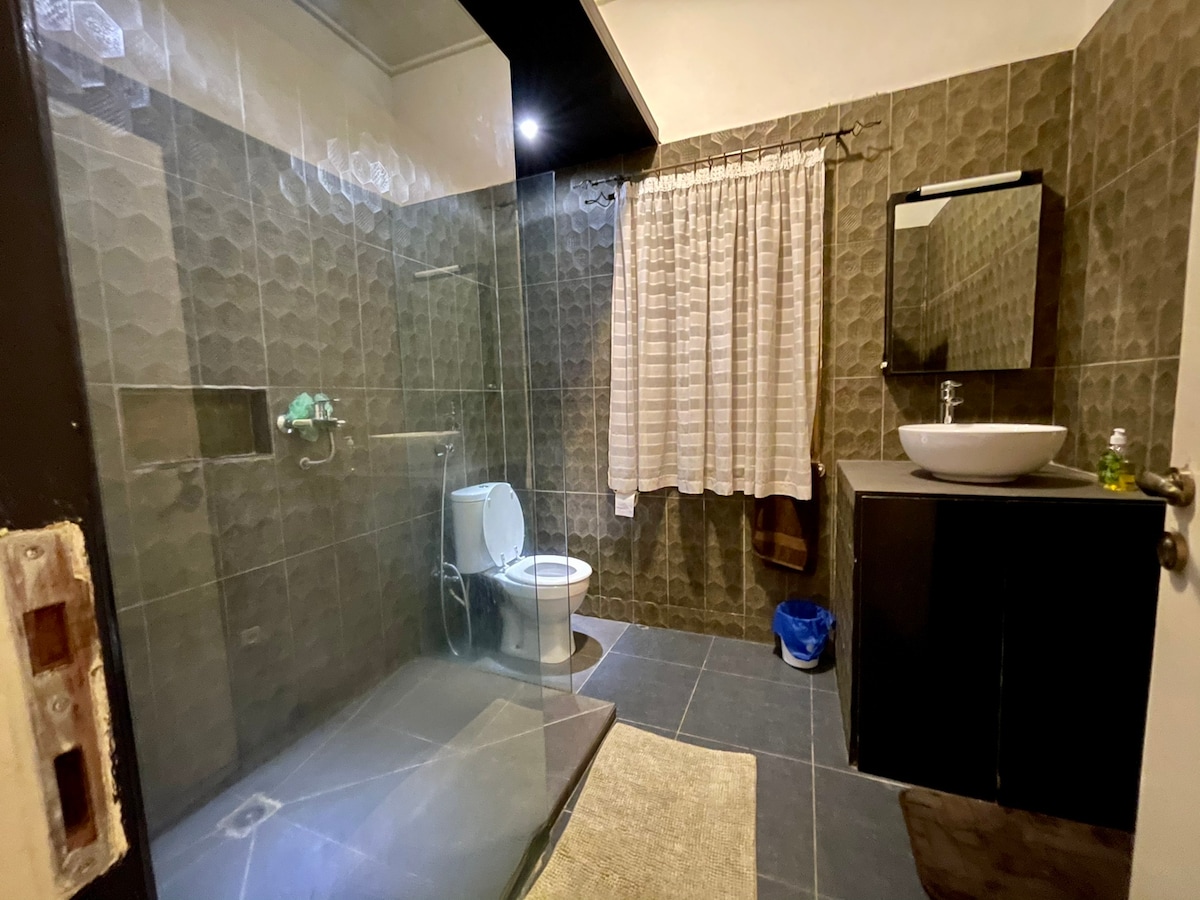 A modern bathroom features a spacious walk-in shower with glass panels and sleek tile work. A minimalistic sink with a round basin is set against a dark vanity. Natural light filters through a partially draped window, complementing the neutral tones of the decor.