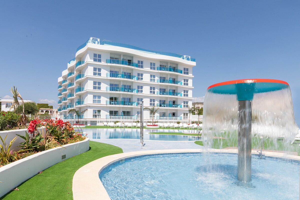 A modern building featuring multiple levels is shown beside a serene pool area. A decorative fountain is visible in the foreground, while vibrant flowers and greenery surround the water features. The structure’s balconies overlook the pool, contributing to a welcoming atmosphere.