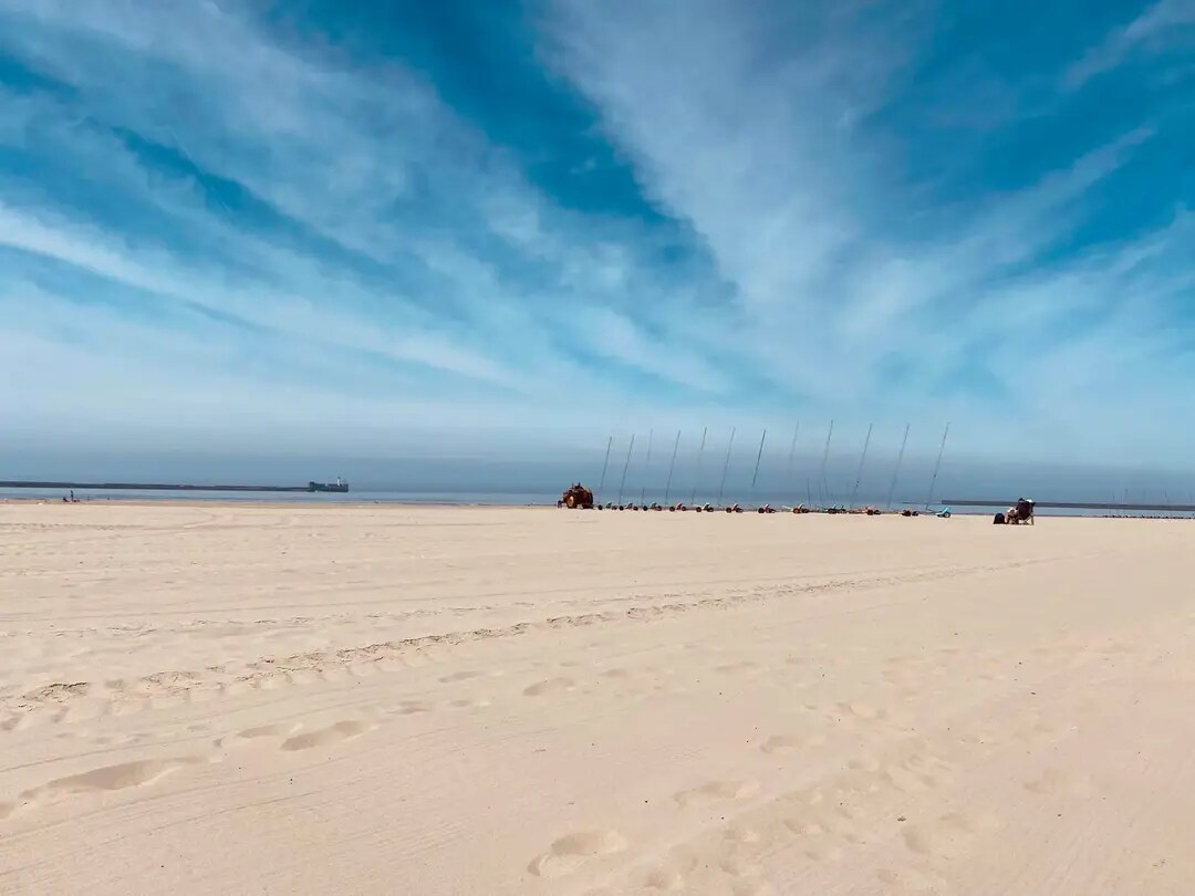 A wide sandy beach stretches under a blue sky filled with soft clouds. Several kite surfers can be seen along the horizon, with tall poles marking their positions. The gentle waves of the sea rise in the distance, creating a serene coastal scene.