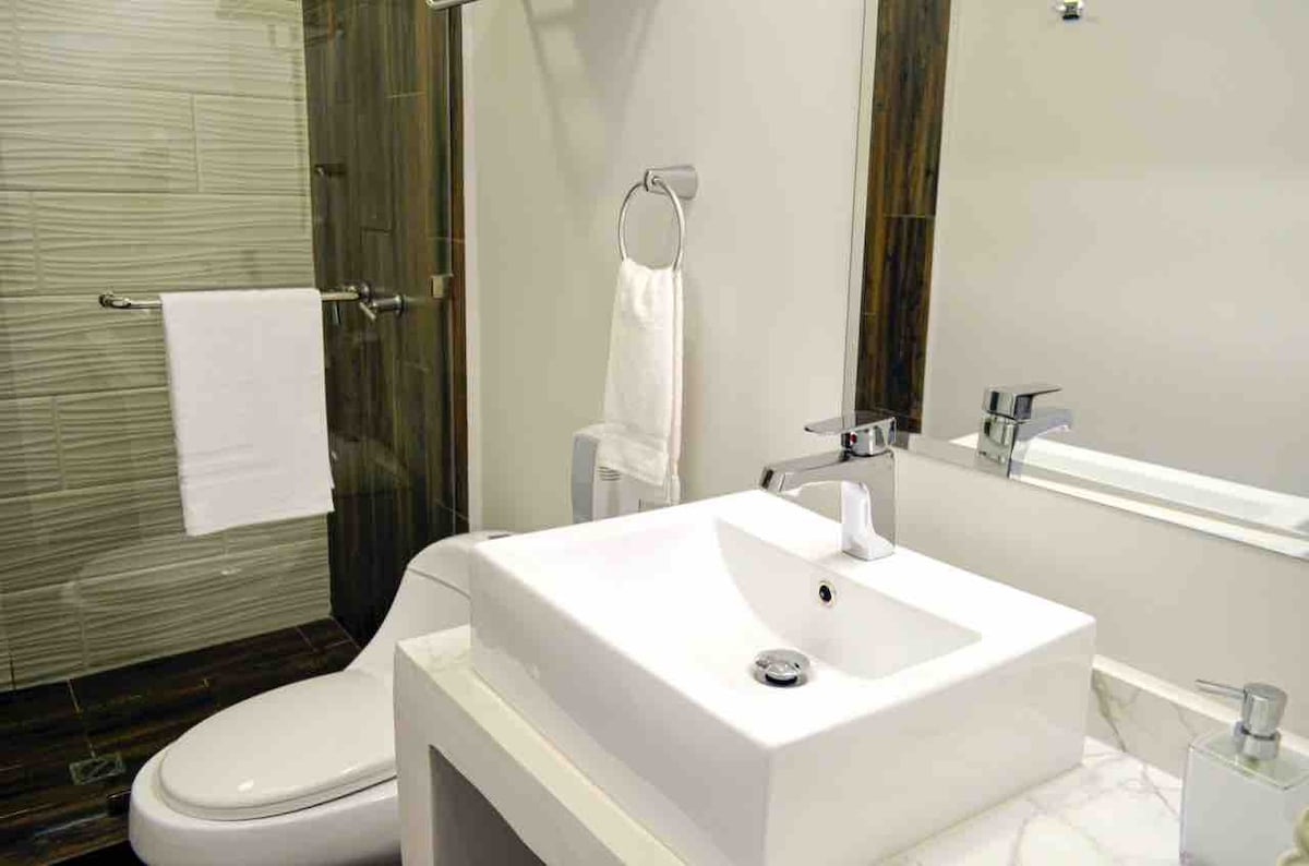 A modern bathroom features a sleek white sink atop a marble countertop, accompanied by a minimalistic faucet. A glass shower enclosure is visible alongside a contemporary toilet. Clean white towels hang neatly on a bar, enhancing the overall tidy appearance of the space.