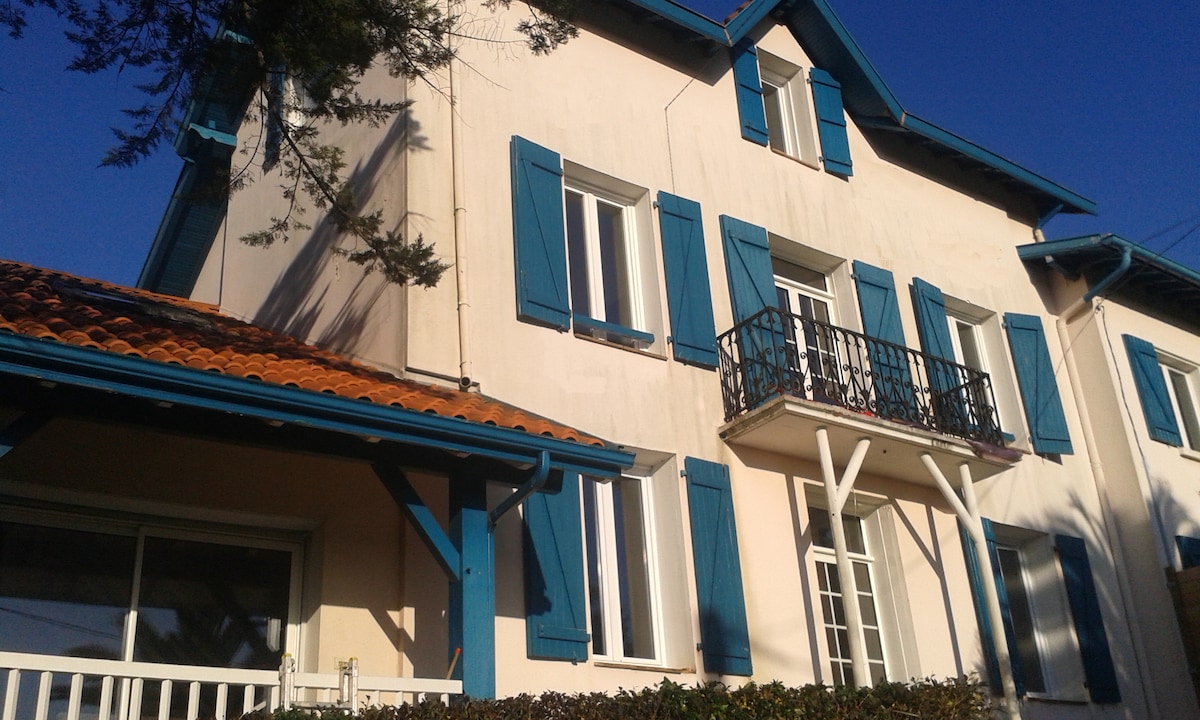 The exterior of a charming house features blue shutters and a terracotta tiled roof. A small balcony is visible on the upper level, offering a view of the surrounding greenery. Large windows reflect the clear sky, contributing to a bright and airy atmosphere.
