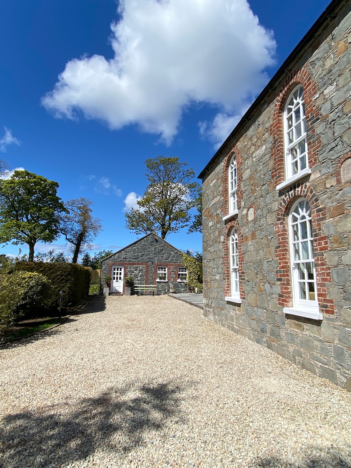 A gravel pathway leads to Ivy Cottage, a stone structure featuring large arched windows. Lush green trees and well-maintained hedges border the pathway, while a clear blue sky with scattered clouds provides a bright backdrop to the serene rural setting.